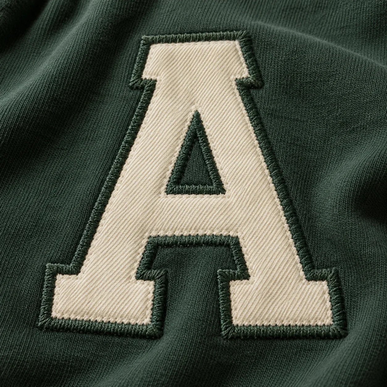 Illustrative close-up of a cream twill letter A with dark green stitched edges on sweatshirt fabric