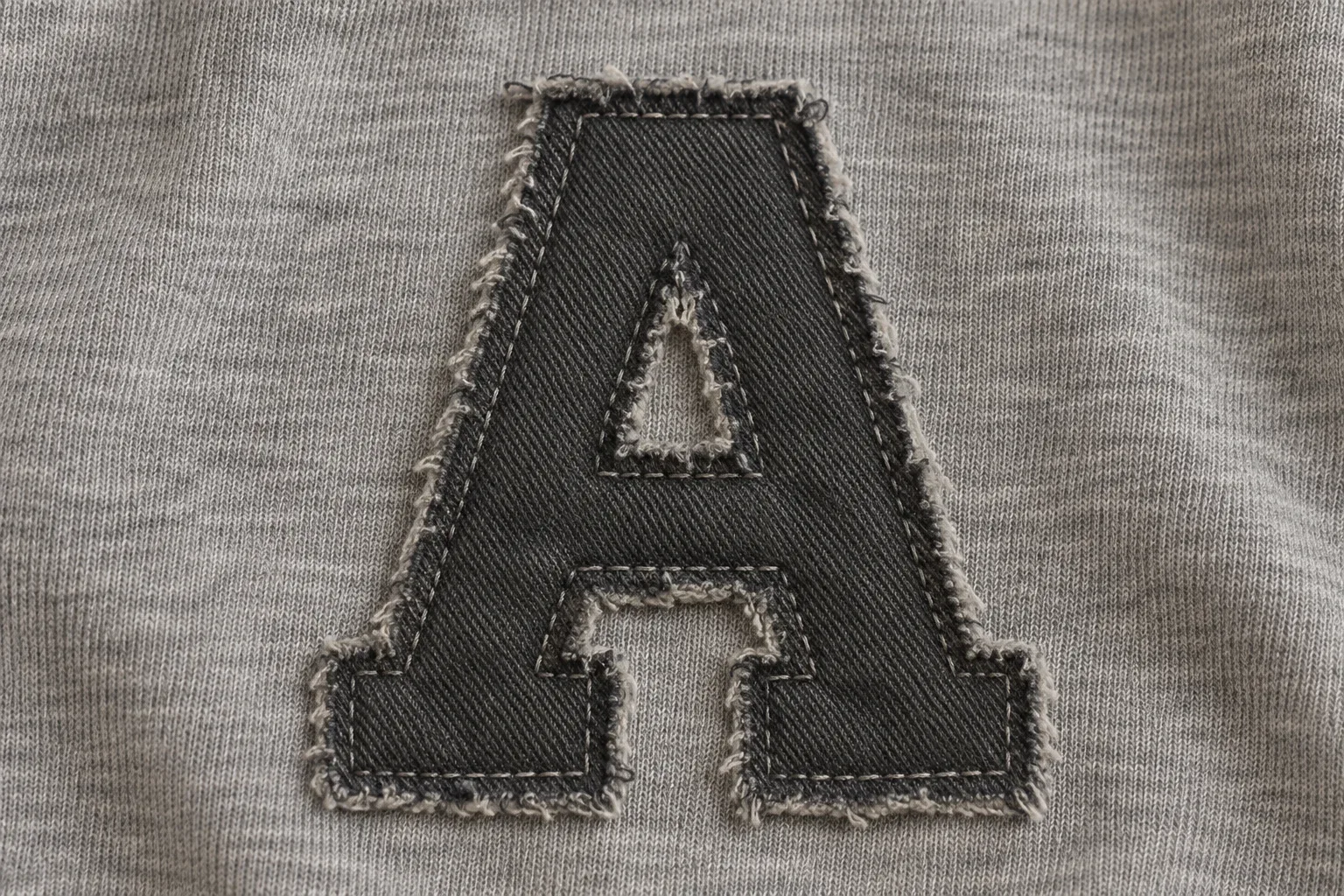 Charcoal fabric letter with exposed frayed edges and a securing stitch line set inside the edge
